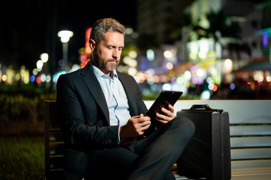 Man in business suit working on tablet outdoors. Business man with tablet outdoor. Business man with a tablet on night street. Businessman using tablet, sit on bench. Night business. - Powered by Adobe