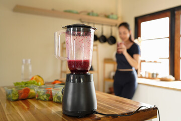 Black electric blender making a smoothie with raspberries, blueberry, with a woman preparing healthy food in the background