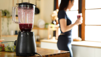 Black electric blender making a smoothie with raspberries, blueberry, with a woman preparing healthy food in the background