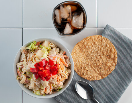 Ensalada de atun con pasta y pimientos rojos encima con una tostada de maiz sobre una servilleta y un vaso con hielo y refresco de cola al lado
