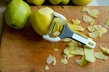 White and gray vegetable peeler with peeled green apples on a brown wooden board. Kitchen utensils for cleaning vegetables and fruits. Healthy eating.