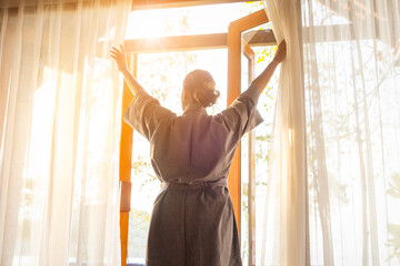 The woman was at the window in the bedroom. She opened the curtains on the window. In the morning and she looks at the view of mountains and trees at sunrise.	