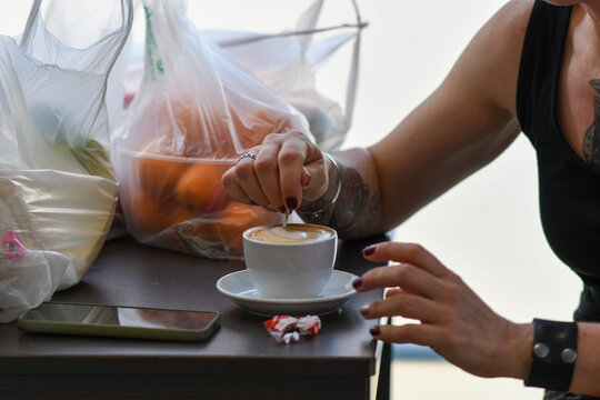 Close up on a woman with tattoos adding sugar to a creamy cappuccino with latte art on a saucer with oranges in the background in plastic bags