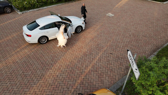 Bride in a white wedding dress getting out of a luxury white car, while the groom in a suit walks towards her on a brick paved driveway with a hotel sign nearby