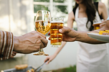 Close-up of friends clinking glasses of beer and wine during an outdoor barbecue party, celebrating and enjoying good times together.