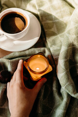 Cozy moment with burning candle in hands, coffee and green plaid in background, autumn or winter mood