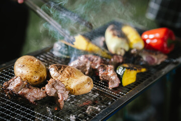 Close-up of grilled meat, potatoes, and vegetables cooking on a barbecue grill with smoke rising, showing delicious outdoor food preparation.