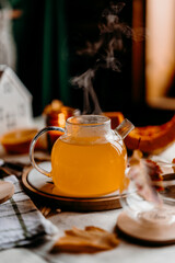 Autumn cozy table scene with transparent teapot of pumpkin tea, candles and leaves.
