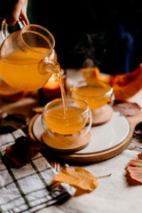 Hot pumpkin tea being poured into glass cups, steam rising in cozy autumn scene with candles and fall leaves.