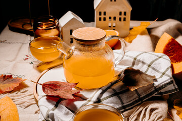 Transparent teapot filled with bright orange pumpkin tea surrounded by candles and autumn leaves. Cozy fall still life with warm steam.