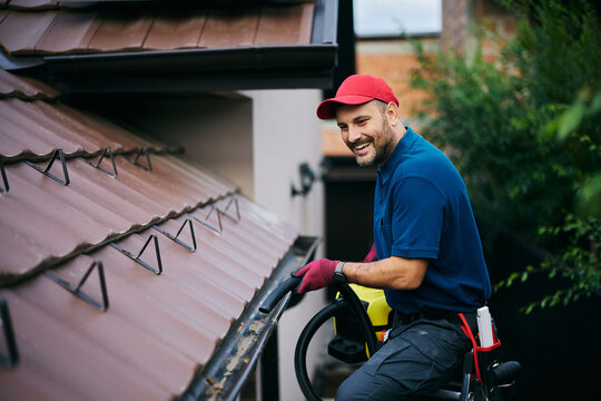 Happy maintenance worker cleaning gutters on roof with blower.