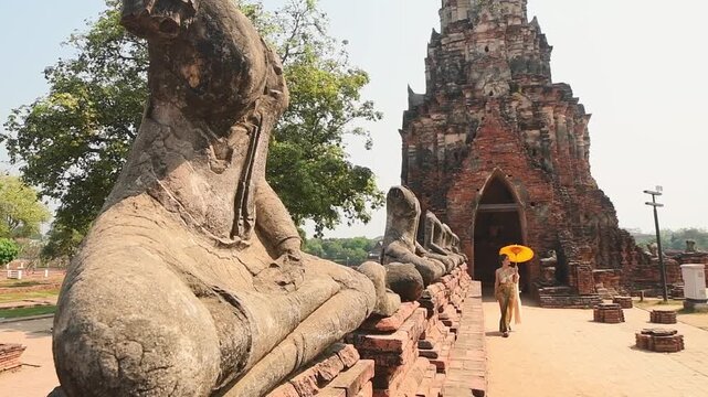 Ayutthaya, Thailand &ndash; March 2023: Woman in traditional Thai dress holding yellow umbrella walking by headless Buddha statues at Wat Chaiwatthanaram, UNESCO World Heritage Site symbol of ancient Siam.