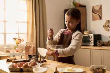 Caucasian girl child whisking ingredients in glass bowl on kitchen counter, preparing food with focused expression, surrounded by baking supplies and utensils in home kitchen