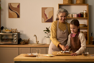 Senior Caucasian woman teaching young Caucasian girl slicing pastry on kitchen counter, both wearing aprons, smiling and interacting while preparing food together indoors