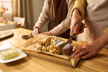 Middle aged Caucasian woman slicing freshly baked cake on wooden board while senior Caucasian woman standing nearby assisting in kitchen, hands visible, butter on plate in foreground
