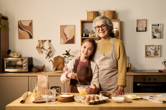 Caucasian senior woman standing beside Caucasian girl mixing ingredients in glass bowl on kitchen counter, both smiling and looking at camera, baking together in home kitchen - Powered by Adobe