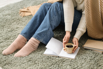 Young woman with cup of hot coffee and book sitting on carpet at home
