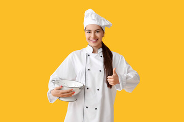 Young female chef with colander showing thumb-up gesture on yellow background