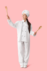 Young female chef with wooden kitchenware on pink background
