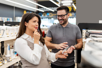 Smiling shop assistant helping a pregnant customer choose home appliances while showcasing various products in a vibrant electronics store