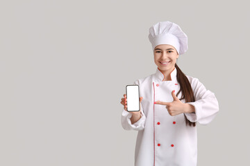 Young female chef pointing at mobile phone on grey background
