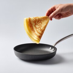 A hand holding a folded crepe over a black frying pan on a white background in a studio shot