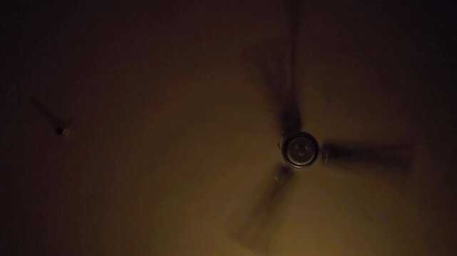 A spinning ceiling fan is visible in a dimly lit room. The fan's motion is blurred. Ceiling fans do not actually cool the air, but rather create a wind-chill effect by circulating air.