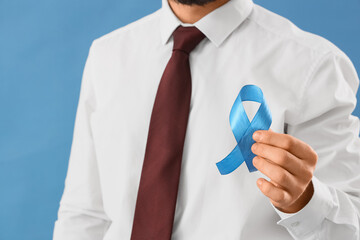 Young businessman with blue ribbon on color background, closeup. Prostate cancer awareness concept