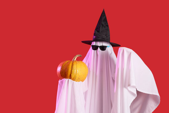 Young man dressed as ghost for Halloween in sunglasses and witch hat with pumpkin on red background