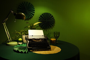 Vintage typewriter, houseplants and glowing lamp on table near green wall