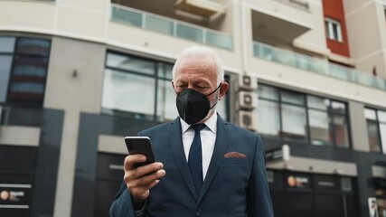 Successful senior businessman wearing a protective face mask walking on a city street and answering a phone call during the coronavirus pandemic, with urban traffic in the background - Powered by Adobe