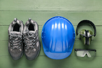 Hardhat, protective earmuffs, safety glasses and boots on green wooden background