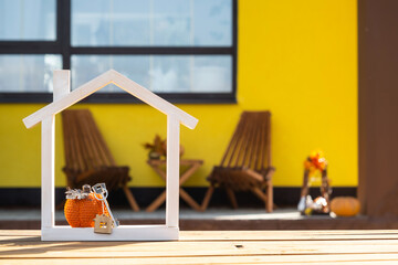 Autumn decor on porch of yellow house outside of pumpkin, lantern, garlands and figure of tiny home and key. Halloween party, autumn mood, Harvest festival, real estate, insurance, mortgage