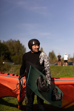 Man in wetsuit holding kiteboard standing in front of orange kite