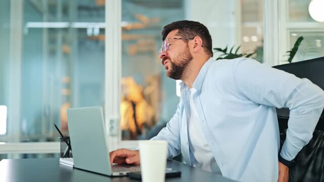 Tired businessman experiences back pain while working on a laptop in a modern office. Stressed male in a blue shirt feeling discomfort from prolonged sitting at desk.