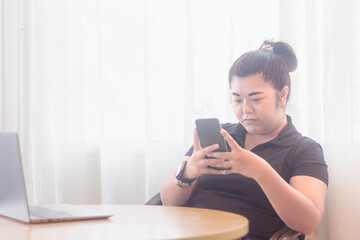 Young woman using smartphone while sitting at a table next to a laptop in a bright, airy room during the day