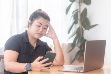 Woman sitting at a table with laptop and smartphone looking concerned in a bright room with a plant