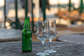 A green glass bottle and two empty glasses rest on a weathered table. The background is softly blurred.