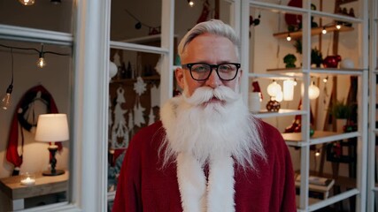 Santa man in red hat stands by festive shelf. Christmas mood fills room with cozy lights. Man smiles in santa hat during holiday. Warm holiday setting with decorated santa man indoors. - Powered by Adobe