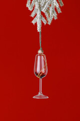Glass of red wine Christmas ornament hanging on fir tree branch on red. New Year glass toy as wineglass with red glitters. Winter holiday aesthetic still life minimal card of celebration, xmas party.