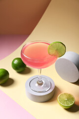 Decorative podium with glass of tasty Cosmopolitan cocktail and fresh limes on colorful background