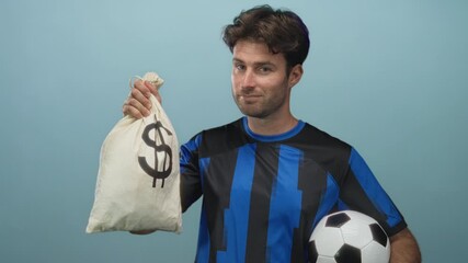 Young man holding money bag and soccer ball in a light blue studio, holding the bag aloft and looking at camera; confidence finance. - Powered by Adobe