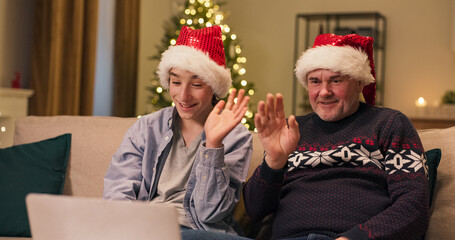 On Christmas Eve, a father and son sit on a couch, having a video call with relatives. They communicate, smile, and wear Christmas hats while a Christmas tree flickers.