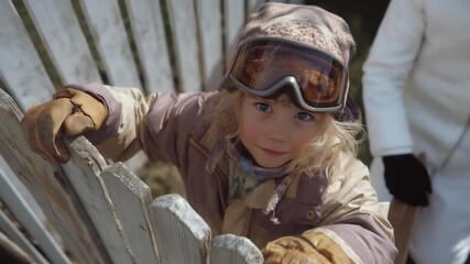 A child, their mittens bulky, tries on skis and goggles in a backyard, the skis' plastic bases slick against trampled grass, bindings loose and unadjusted, and the goggles' strap tangled around