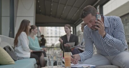 Diverse business professionals hold an informal team meeting on a modern outdoor terrace. A focused man takes notes while talking on his phone, symbolizing flexible work and dynamic collaboration. - Powered by Adobe