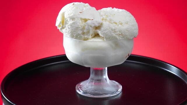 Scoops of vanilla ice cream in a glass bowl slowly rotate against a brightly colored background.