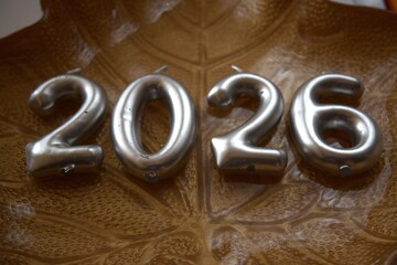 Silver colored number candles arranged as 2026 on a decorative plate with leaf pattern. Symbol of calendar date and new beginnings.
