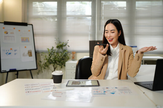 Asian businesswoman in a beige suit talking to voice assistant or chatbot on her smartphone in office, sitting in modern office, using ai technology, smart assistant.