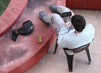 Perspectiva aérea de chico joven sentado mirando un vídeo en su teléfono móvil con cigarro en la mano, taza de café y riñonera en el patio 
