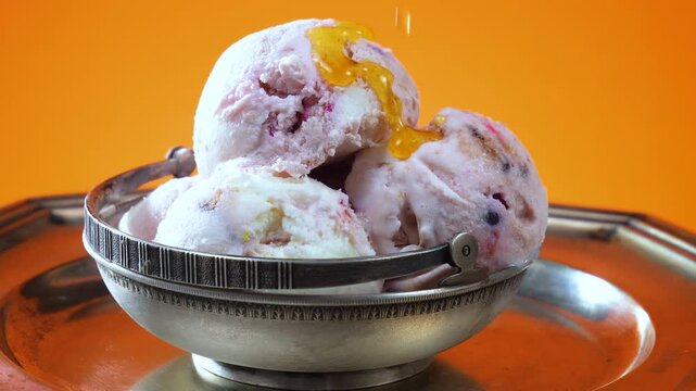 Balls of chocolate ice cream in a metal bowl, drizzled with fruit syrup, slowly rotate against a brightly colored background.